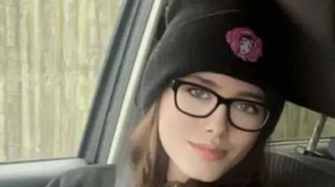 Facebook A head and shoulders image of Jamie-Lea Biscoe sitting in the front seat of car. She has long brown hair and is wearing black framed spectacles and a black beanie. Her face is turned towards the camera, with the side window behind her. 