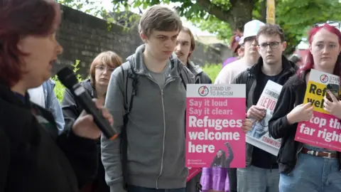 A group of protestors outside the new councils' first meeting, some holding placards. One of the placards reads: "Say it loud, say it clear: Refugees welcome here"