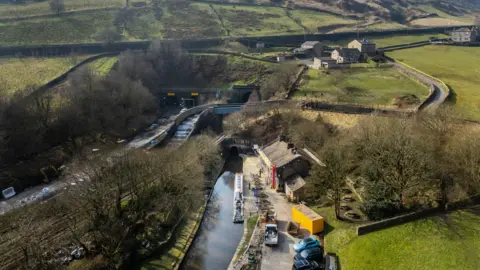 PA Media A quiet village scene with a canal running through it and a tunnel entrance set into a hillside. Stone buildings, green fields and a narrowboat create a peaceful countryside setting.