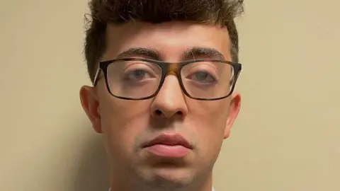 PA Office of Attorney General Anthony Abbatiello stares straight at the camera in his booking photo, wearing glasses and curly brown hair