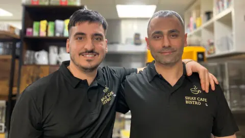 Elizabeth Baines / BBC Dos hombres visten polos negros. Detrás, se ve borrosa una cocina.