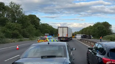 A lorry is seen on its side on the carriageway on the right on the image. The carriageway on the left, which is more prominent on the photo, includes an upright lorry, a police car, a trafffic cone on the left and two other cars at the bottom of the image.