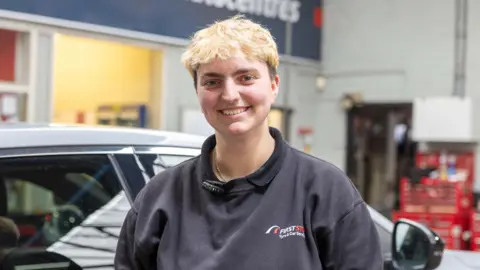 A woman with mainly blonde hair and wearing a dark top is smiling at the camera. She is next to a silver car and a sign above her head in the background includes the letters CC Autocentres.