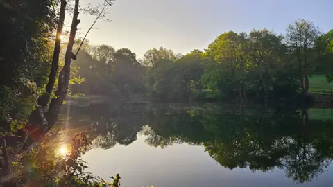 Stormchaser Al / BBC Weather Watchers Sunrise over a lake in Halesowen. The sun reflects on the water to the left, with trees lining the bank and a light early morning mist in the air
