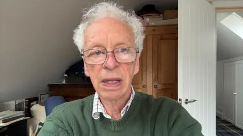 David Adams A man with grey hair and glasses, wearing a green jumper and a checked shirt underneath pictured from the shoulders up with a wardrobe, a chest of drawers and a door behind him.