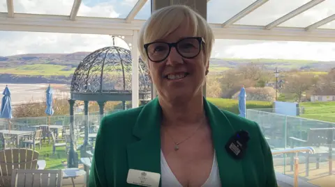 Beverly Rutherford, business development manager at The Victoria Hotel. She has short blond hair and is wearing glasses with a black rim, a white v-neck t-shirt and green blazer. She is standing in front of a window, with a terrace behind her, looking over Robin Hood's Bay.