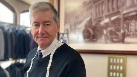 Neil Warwick, standing in a shop with a tape-measure around his neck. He has grey hair and is wearing a white shirt, blue tie and a navy V-neck jumper.