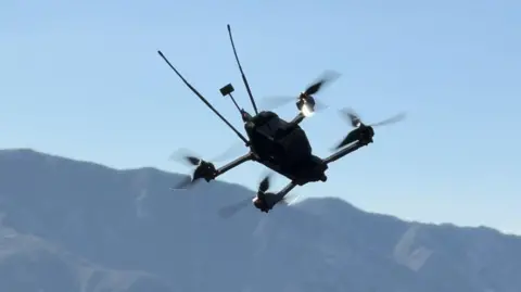Image of a drone with four propellers flying over a mountainous terrain. In the background can be seen misty hills and a blue grey sky.