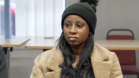 Tilisha Goupall wearing a coat and black hat sitting in a room infront of same tables and a chair 