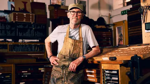 Nick Hand wearing a grey t-shirt and brown apron, which is heavily stained with black ink. He is leaning on a wooden table inside his workshop, looking straight at the camera with his hand on his hip.