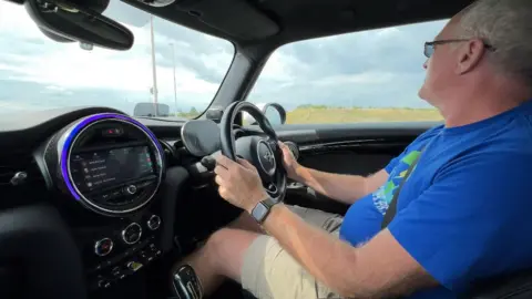 Mike Nunn is driving his electric Mini. He has two hands on the steering wheel and is focused, facing forwards and looking directly out of the front window. He is wearing a blue t-shirt and beige shorts. He is also wearing sunglasses.