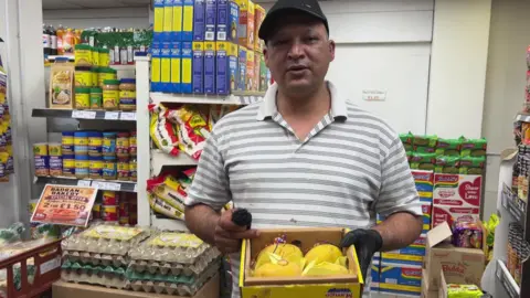 Aisha Iqbal/BBC A man stands in the middle of a shop , surrounded by shelves of foodstuffs. He is holding a box of ripe, yellow mangoes. He wears a black cap and a striped shirt.