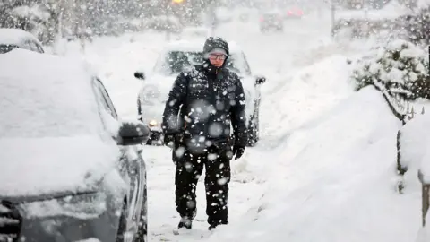 A man walks along a heavily snow covered street with his hood up.