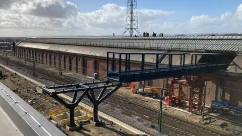 Network Rail A black footbridge is being installed in sections over a single railway line at Darlington Station. Behind the footbridge is the long red-brick building which makes up part of the original station building.