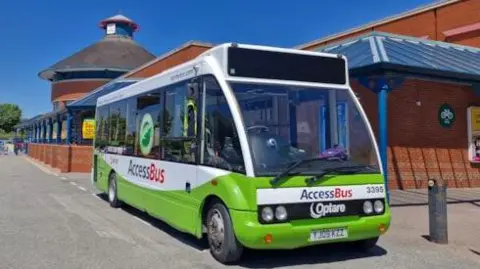 Green West Yorkshire AccessBus parked outside a red brick bus station, with bright blue sky above