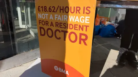 A banner, which is yellow, white and orange, including £18.62/hour is not a fair wage for a resident doctor is prominent in the middle of the image. 
