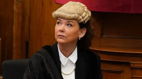 Dorothy Bain, who has long dark hair tied back under a court wig, looks up to her right while sitting down. She is wearing a black gown over a white shirt, with wood panelling behind her. 