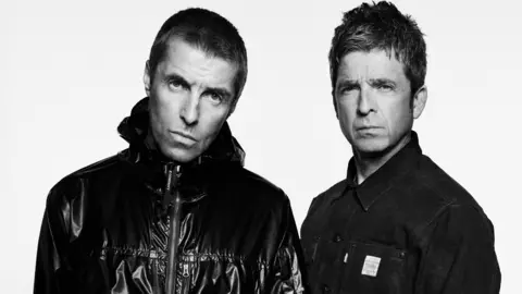 Black and white shot of Liam Gallagher (L) with short cropped hair and black bomber jacket and elder brother Noel (R) with short spiky hair and a dark denim shirt.