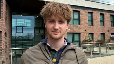 A young man with light brown hair and light stubble stands in front of a red-brick building. He wears a navy blue rugby style shirt and a khaki green coat.