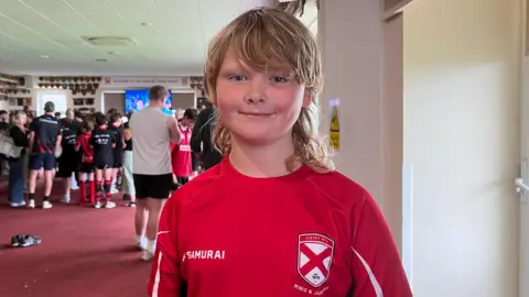Bay-Zander is wearing a red Jersey RFC Minis and Juniors sports jersey as he stands in the foreground of a clubhouse. Visible text on the shirt includes 'SAMURAI' and 'Santander International'. In the background, a group of people and young sports participants are gathered near a television screen in a large room with red carpet and walls decorated with plaques and awards.