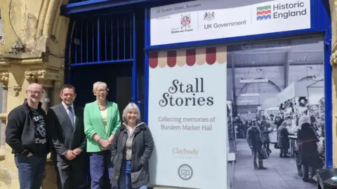 Four people stand in front of a building with a large sign that reads "Stall Stories: Collecting memories of Burslem Market Hall." The sign says the project funded by the City of Stoke-on-Trent and the UK Government, with Historic England also recognised.
