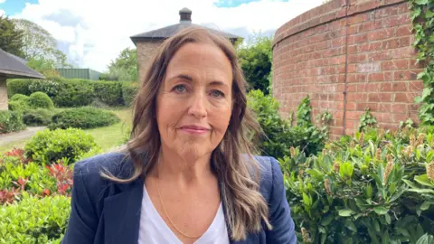 Jo Monk photographed in a garden. She has long wavy brown hair and is wearing a white top and a blue jacket.