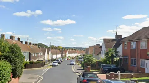 A residential street that curves to the right in the distance with semi-detached houses on a sunny day, with small white clouds in the sky. There are cars parked along the road in the residential area, and a T-junction with another road can be seen. 