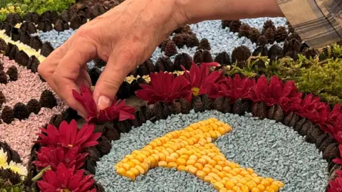 A hand is pressing a petal into a design on a wooden board. The board contains petals, sweetcorn, moss, painted rocks and beech nuts.