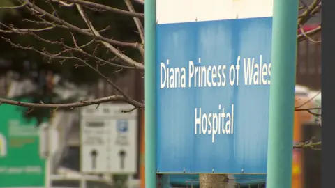 The photo is focussed upon a blue and white sign. It reads 'Diana Princess of Wales Hospital.' Though the background is blurred, there are other signs in the distance signposting for the car park. 
