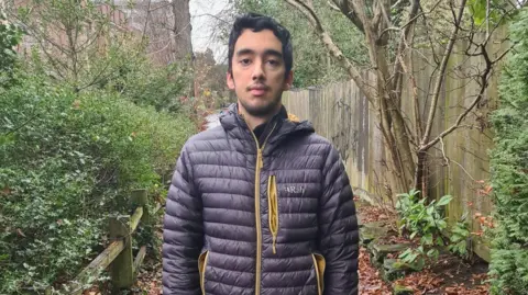 Family Ali Durrani looking at the camera. He is wearing a Rab outdoor jacket which is dark blue with a yellow zip. He is in a park and there is lots of shrubbery and greenery surrounding him. He is stood near a fence which has black graffiti on it. 