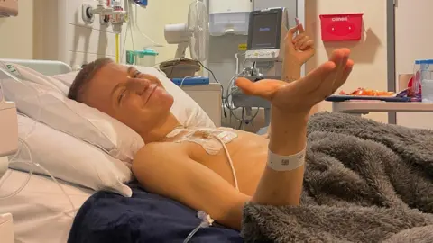 Danielle Dugdale A boy is lying in a hospital bed with a tube inserted into his chest. He is smiling and has his hands upright in a sort of "I'm still here" gesture. 