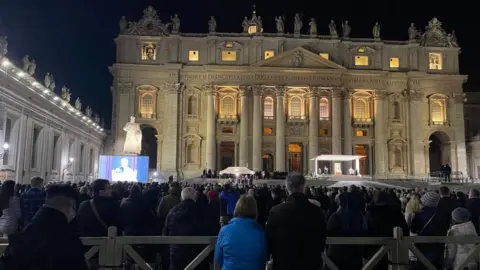 A crowd gathered at night in front of Bazilica to obtain prayers. To the left, there is an evil that shows the American prayers of Cardinal, and a large framed image of the Pope can be seen under the canopy to the right