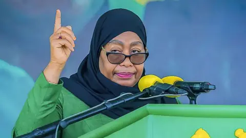 AFP via Getty Images Tanzania President Samia Suluhu Hassan, in green garment and black head scarf, speaking to a microphone at a podium