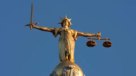 A gold-coloured statue of Lady Justice stands on top of a domed building against a clear blue sky. She is holding a sword in one outstretched hand and a set of scales in the other.