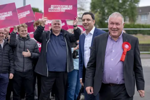 PA Media Davy Russell in a suit and red rosette with Anas Sarwar and a crowd behind him holding up placards