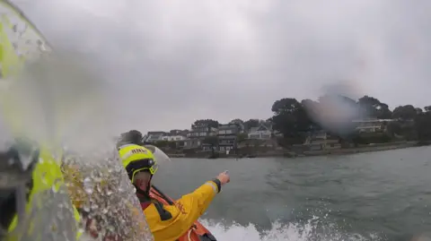 View from a lifeboat crew's camera. Their colleague is pointing towards the shore on a grey, rainy day.