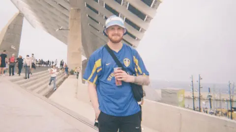 Kris Smith A man is stood on some concrete steps. He is wearing a blue Leeds United shirt and a bucket hat. He is holding a paper cup.