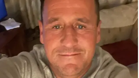 A selfie image showing a man in his 40s with receding brown hair. He is in a living room with a sofa and the corner of a TV visible. He is wearing a light grey top.