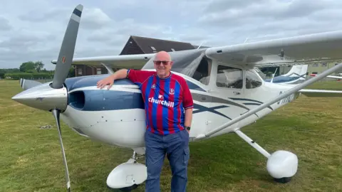 He stands beside a small single-engine plane on a grassy airfield, wearing a red and blue striped team shirt and leaning on the aircraft’s nose.