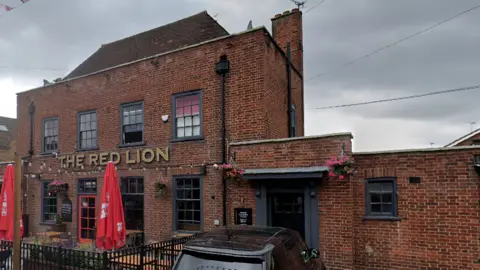 The outside of the Red Lion pub, a large brick building with a beer garden. A black car is to the left and a woman is walking past, wearing a blue top and blue trousers.

