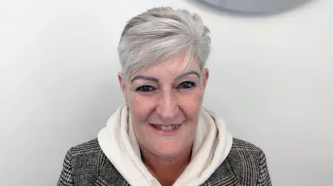 A head and shoulders crop of Val Lucus in a checked jacket and white hoody smiling and looking into the camera against a grey background.