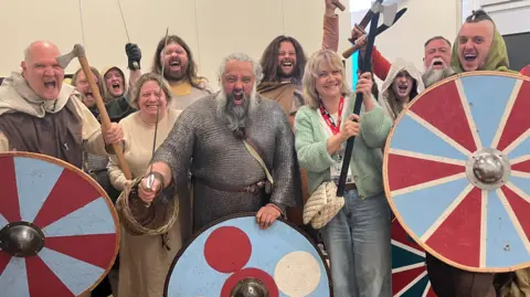 A group of 11 men and women dressed as Vikings, including a man in chain-mail armour and some holding shields and swords, cheer as they stand in a tent. 