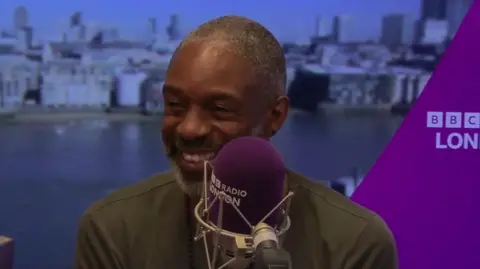 Wil Johnson in a BBC London radio studio. He wears a green t-shirt and sits behind a purple microphone.