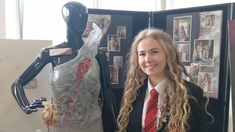 A girl is smiling next to a mannequin. She has long, curly, blond hair. She is wearing her school uniform.
