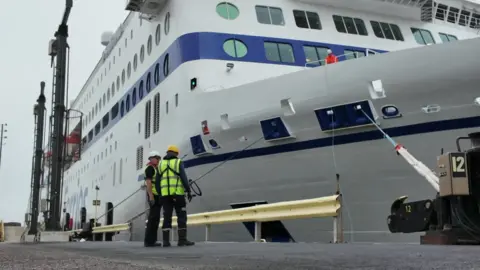 A large ferry pulls into port. Two men in high-vis are standing dockside.