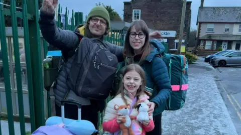Marratravels Craig, Summer and Alli Henry outside Corkickle station. They are standing outside a green metal fence, with the station sign visible in the background. They are carrying rucksacks and bags and are wrapped up in winter coats. Craig is weaving with one arm. Summer is holding two teddy bears. Alli is behind her and has her hand on Summer's shoulder. They are all smiling.