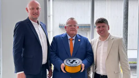 Three men in suit jackets look into the camera. The men on the left is tall and bald-headed with a blue jacket and white shirt. The man in the middle is shorter, with receding hair and glasses and is in a blue jacket with a light blue shirt and orange tie. He is holding a reel of orange fibre. The man on the right has short, curly hair in a grey jacket with a white shirt.