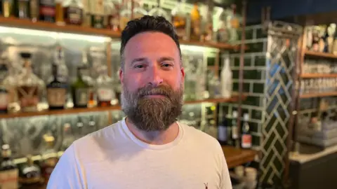 The image shows a man standing behind a bar in a cocktail bar in Newquay. He has a beard and is wearing a white t shirt. There are rows of bottles on the shelves behind him. 