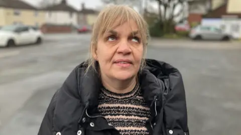 A woman with blonde hair, wearing a black jacket over a stripy style jumper. She is the focus of the picture, with cars and houses blurred in the background.