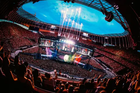HYBE / Big Hit Music A wide shot of BTS performing in London, with thousands of fans watching as confetti and fireworks shoot into the sky